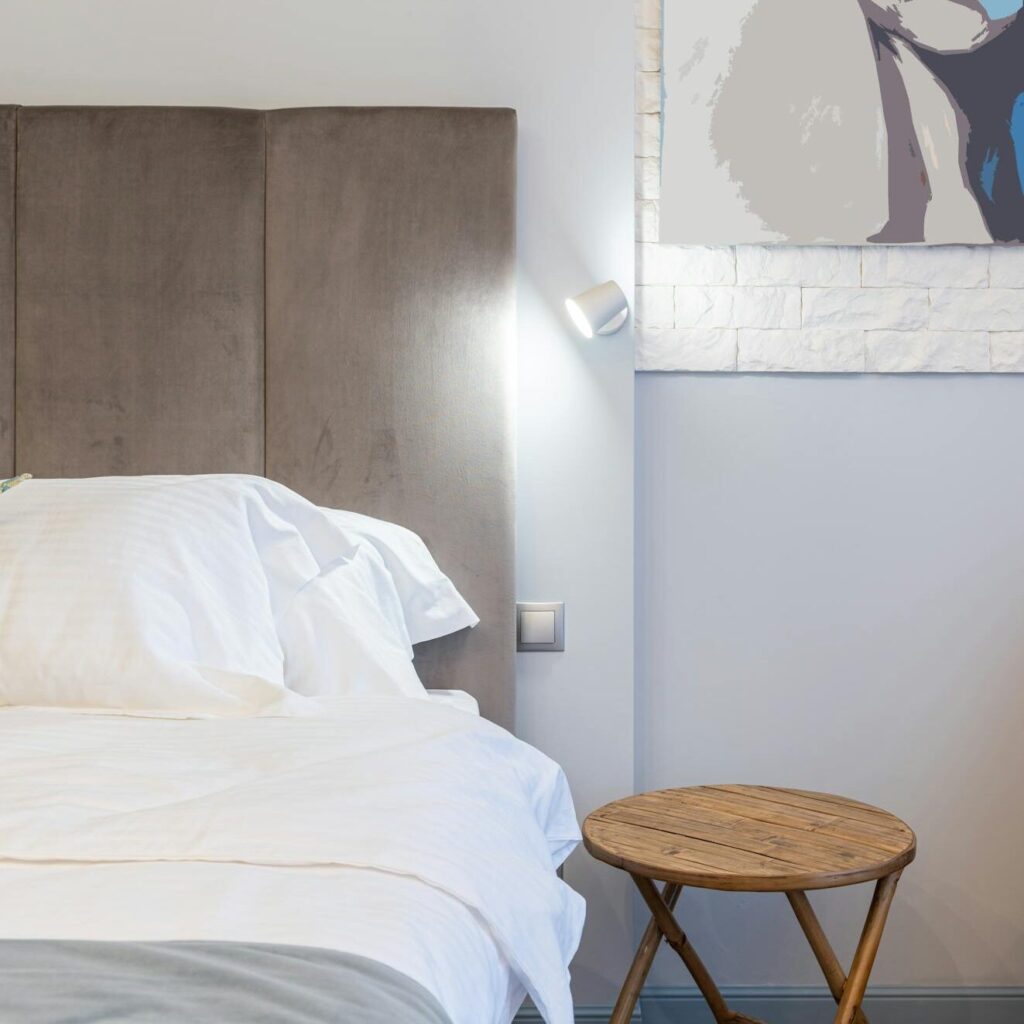 Contemporary bedroom with crumpled cover on bed illuminated by artificial light in house with folding table and painting on wall